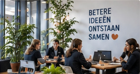 Mensen in overleg rond een tafel in een lichte werkruimte met planten, met aan de wand een spandoek met de tekst betere ideeën ontstaan samen.