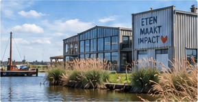 Bedrijfsgebouw met veel glas aan het water, met riet langs de oever en een bord met de tekst eten maakt impact.
